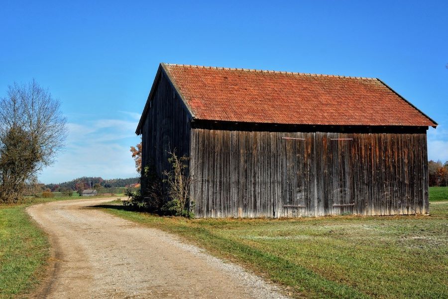 A wood barn with a red roof in the countryside next to a small gravel and dirt road.