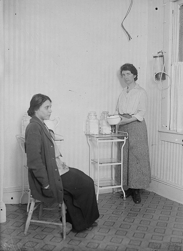 Fania Mindell sits in a chair facing to the side. Margaret Sanger stands next to a side table containing jars of towels, holding a bowl and facing the camera.