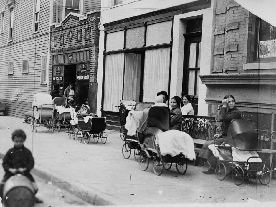 Numerous strollers are parked on a sidewalk outside of a building. A few women sit on a bench. A child plays in the street.