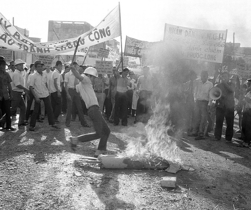 A banner-waving crowd is gathered around a man who is kicking an effigy burning on the ground.
