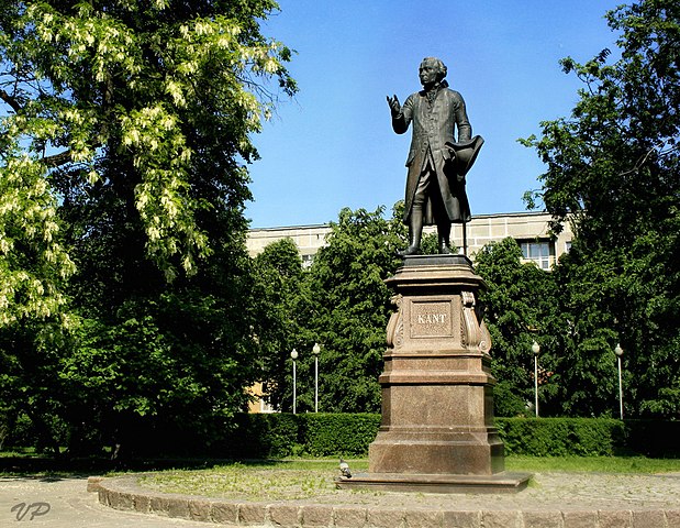 A statute in a park in Konisberg of a man in 18th century clothing on a pedestal with 'KANT' on the front.