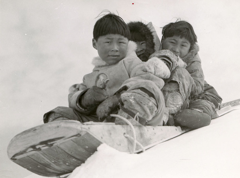 Three children wearing winter furs and riding a sled.