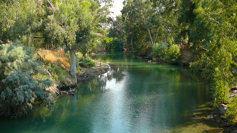 A bend in the Jordan River with green, gently rippling water surrounded by much vegetation.