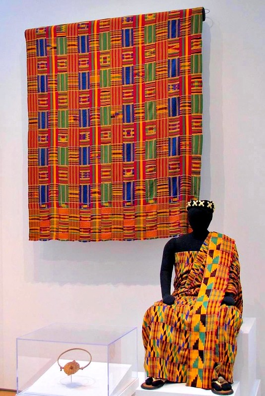 A colorful robe is worn by a seated dummy while a similarly colorful tapestry hangs on the wall behind it.