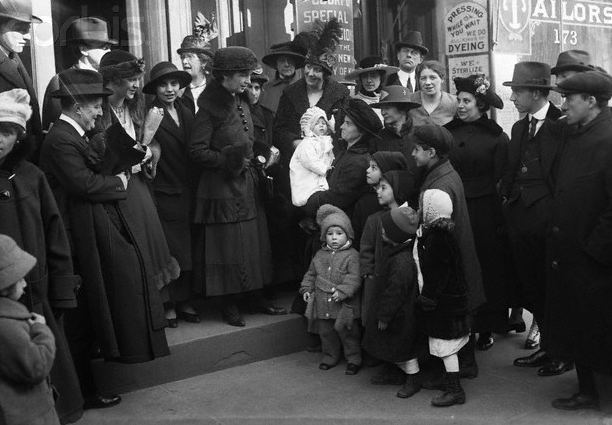 Numerous men, women, and children in overcoats and formal attire surround Margaret Sanger and Fania Mindell as they stand on the steps to a building.