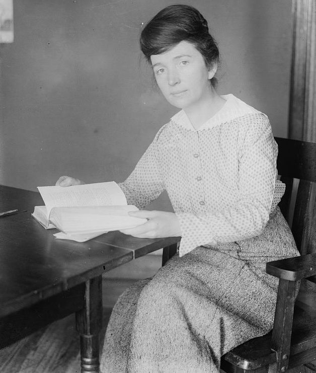 Margaret Sanger seated at a table and holding an open book.