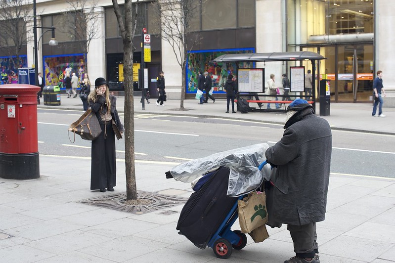 A woman in nice clothes and carrying a designer handbag talks on a mobile phone while a homeless man pushes a plastic covered dolly along the same sidewalk.