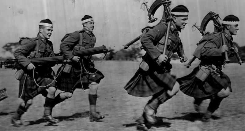 Four men in kilts and military uniforms run while carrying machine guns.