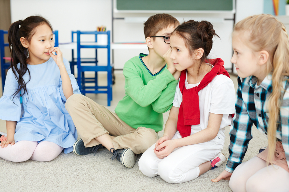 Four children in a classroom, with one child whispering into the ear of another.